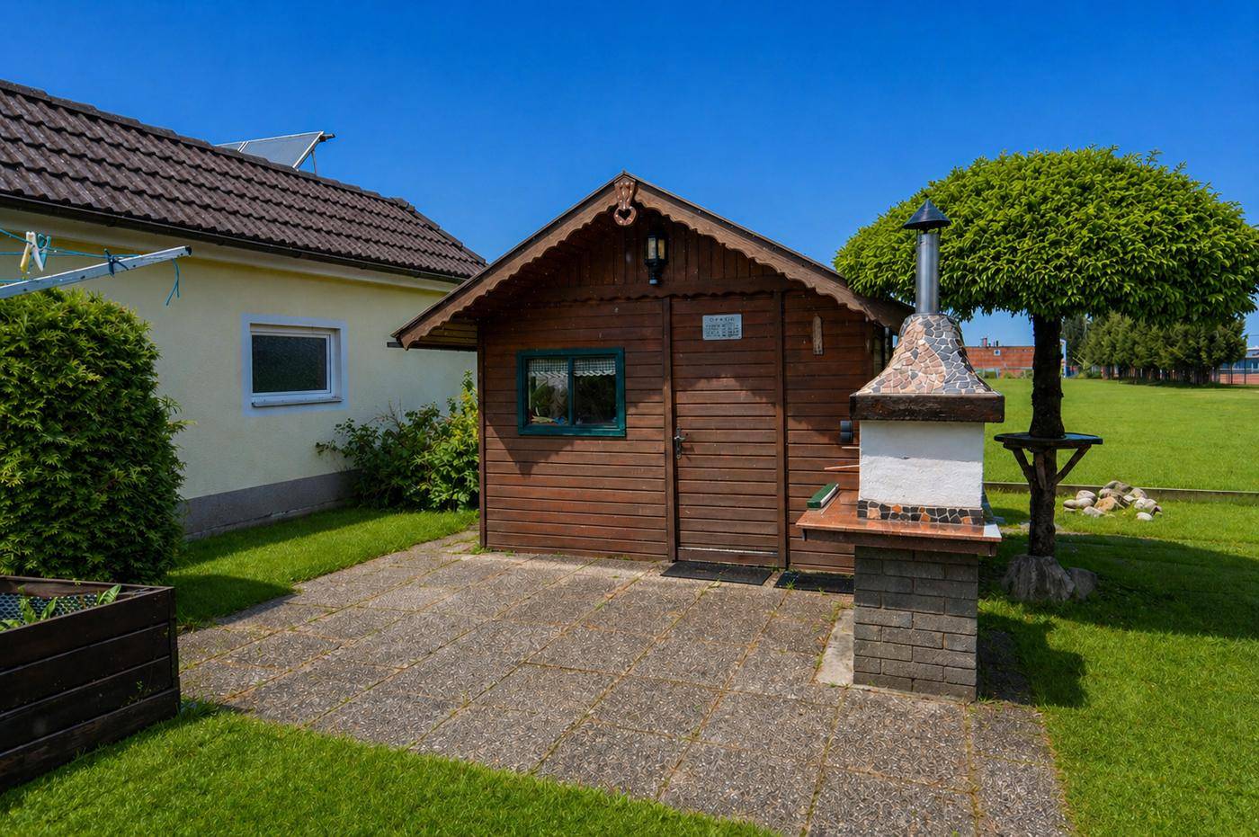 Gartenbereich mit einem Holzschuppen, gepflastertem Grillplatz und Rasenfläche.