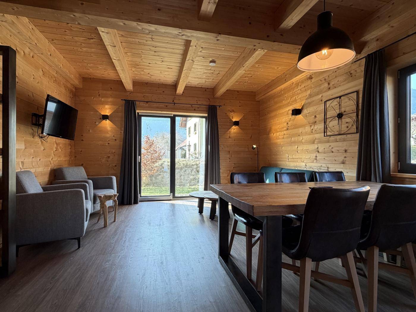 Gemütlicher Wohn-Essbereich mit Holzwänden, Holzbalkendecke und Zugang zum Balkon.