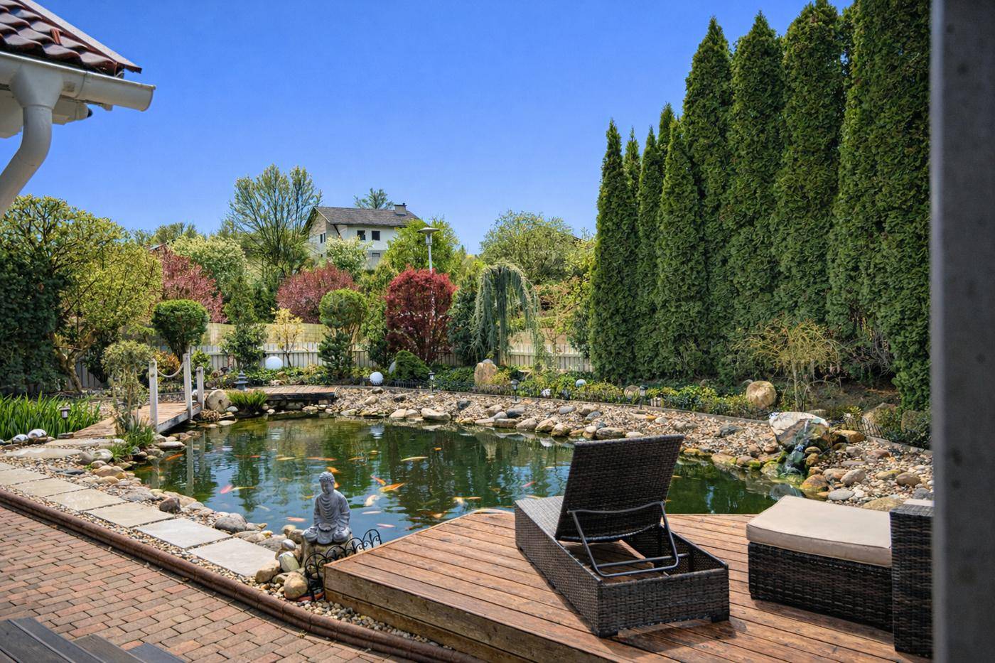 Entspannungsbereich im Garten mit großem Teich, Holzdeck und Liegestühlen unter blauem Himmel.