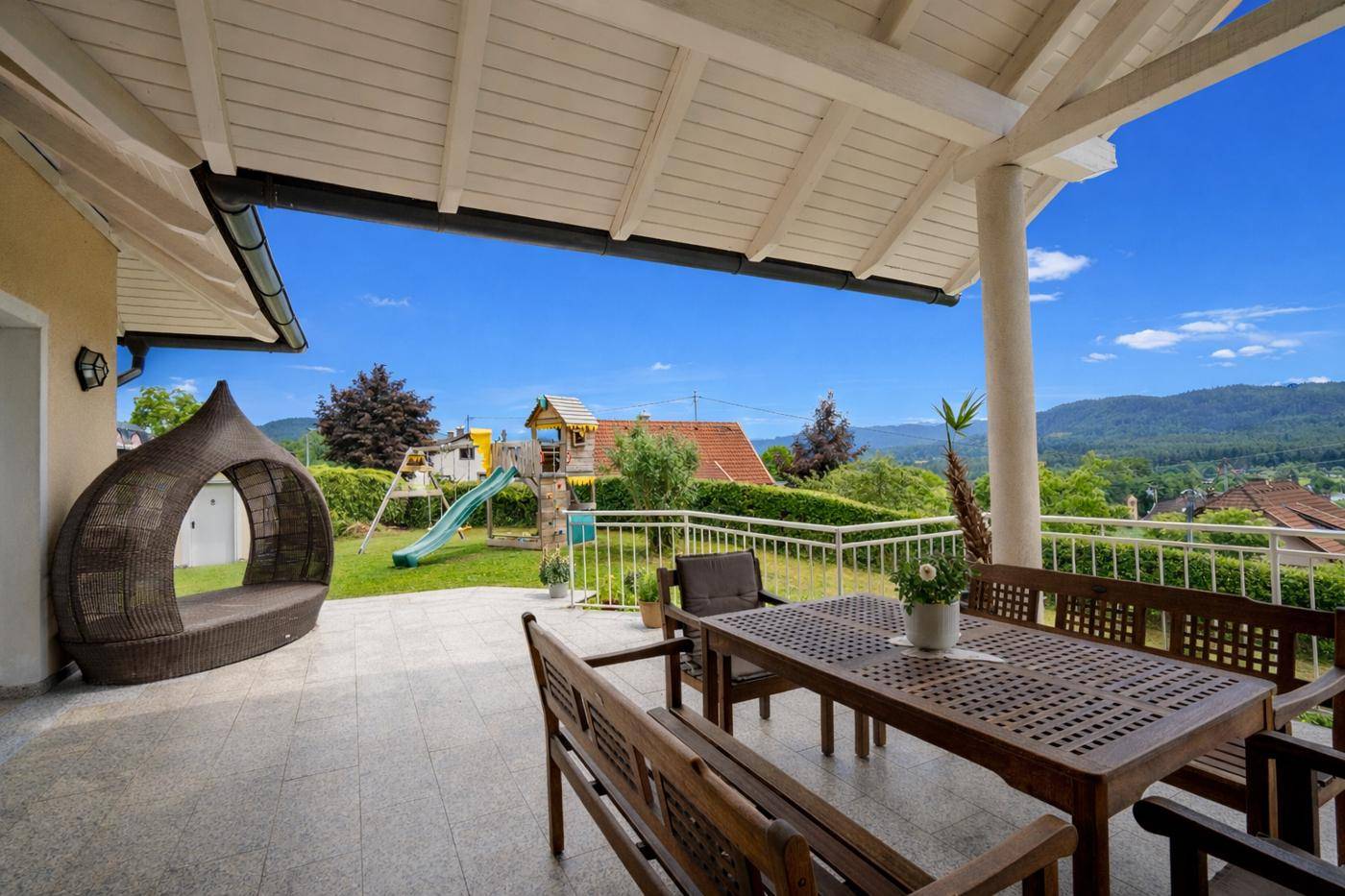 Überdachte Terrasse mit Gartenmöbeln, Blick auf die Berge und einen Kinderspielplatz im Garten.