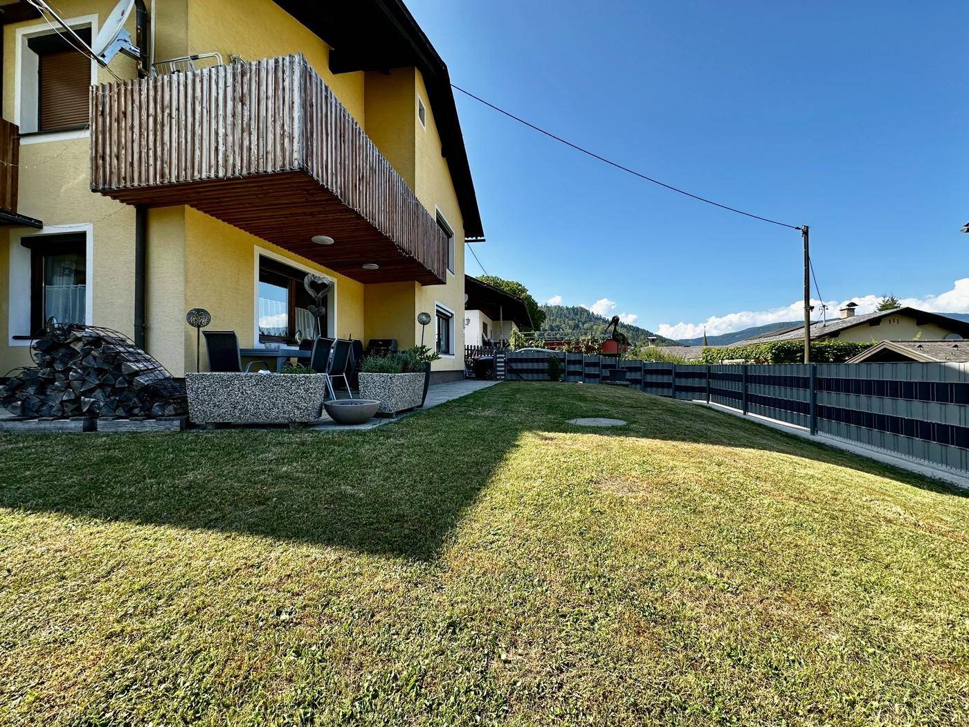 Gelbes Haus mit Balkon, Terrasse und gepflegtem Rasen, umgeben von einer Hecke und Holzstapeln.