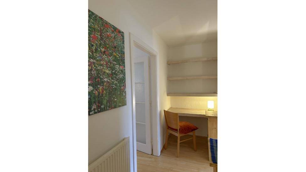 Kleiner Arbeitsbereich mit Schreibtisch, Regal und einem farbenfrohen Gemälde an der Wand.