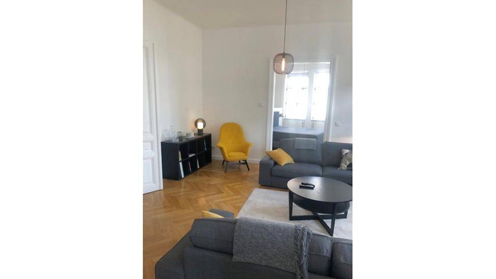 Heller Wohnbereich mit grauem Sofa, gelbem Sessel und Parkettboden, offen zu einem weiteren Raum.