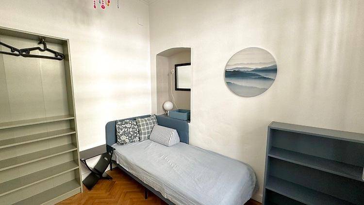 Kleines Schlafzimmer mit Einzelbett, Regal und moderner Wanddekoration in hellem Ambiente.