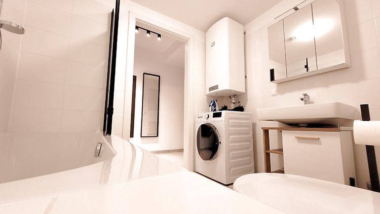 Modernes Badezimmer mit integrierter Waschmaschine, Dusche, Waschbecken und Spiegelschrank in hellen Farbtönen.