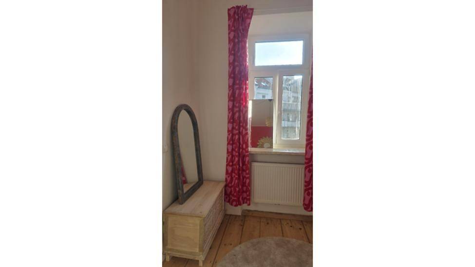 Schlafzimmer mit Holzboden, einem Fenster mit roten Vorhängen und einem großen Spiegel.