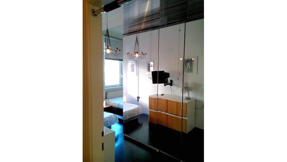 Schlafzimmer mit Spiegelwand und Fernseher, durch eine Glaswand vom Flur getrennt.