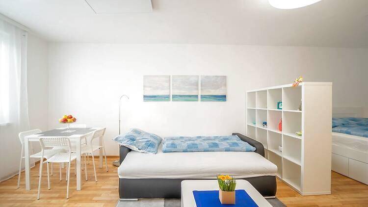 Helles Wohnzimmer mit Sofa, Esstisch und Regal, Teil eines Studio-Apartments mit Schlafbereich.