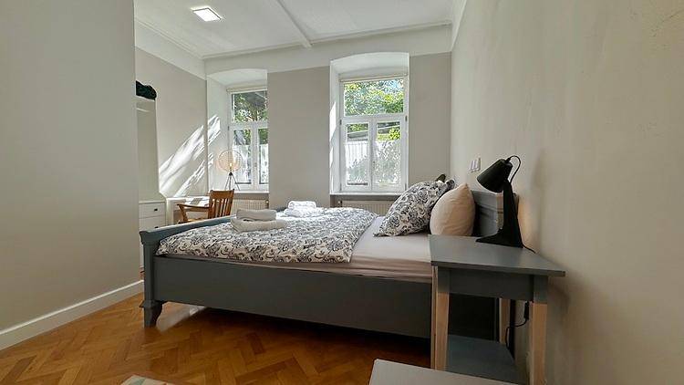 Helles Schlafzimmer mit zwei Fenstern, Doppelbett und Holzboden, gut beleuchtet mit Grünblick.