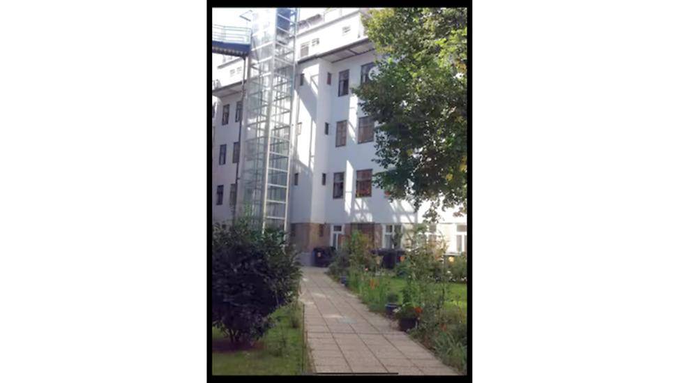Außenansicht eines Gebäudes mit einem modernen Glasaufzug und einem gepflegten Gartenweg.