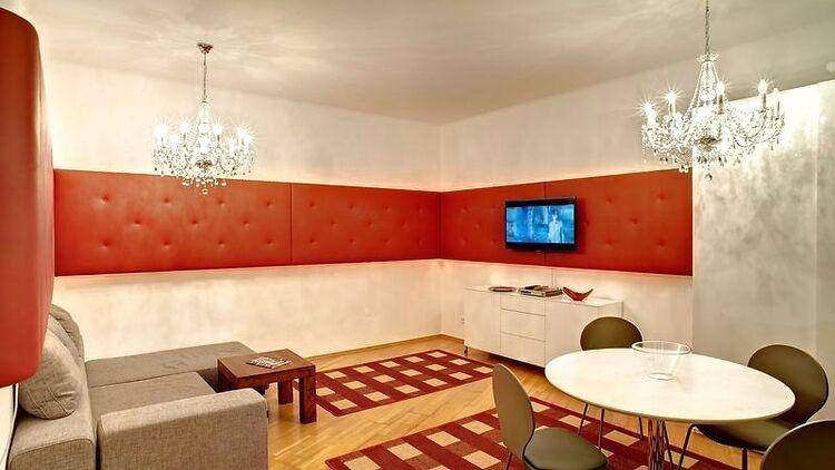 Helles Wohn-Esszimmer mit roter Akzentwand, bequemer Couch und glitzerndem Kronleuchter.