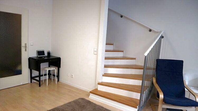 Heller Wohnbereich mit Holztreppe, kleinem Schreibtisch und Zugang zu einem weiteren Raum.