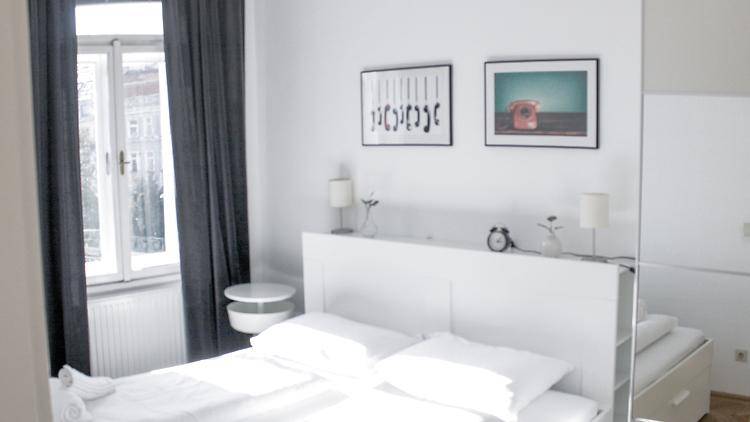 Freundliches Schlafzimmer mit weißem Bett, minimalistischer Einrichtung und viel Tageslicht durch das Fenster.