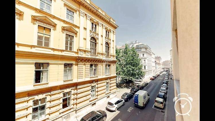 Historische Gebäudefassade an einer belebten Straße mit parkenden Autos und Bäumen.
