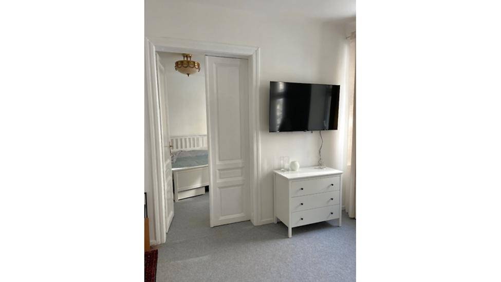 Wohnbereich mit Wandfernseher und Kommode, der einen Durchgang zum Schlafzimmer bietet.