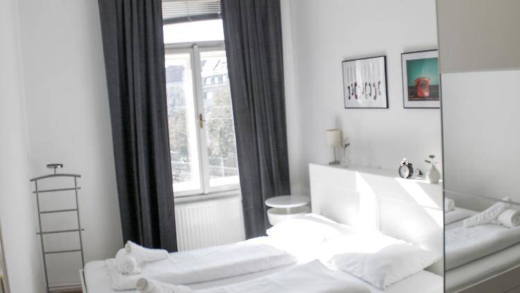 Helles Schlafzimmer mit weißem Doppelbett, Nachttischen und dunklen Vorhängen, gut belichtet durch das Fenster.