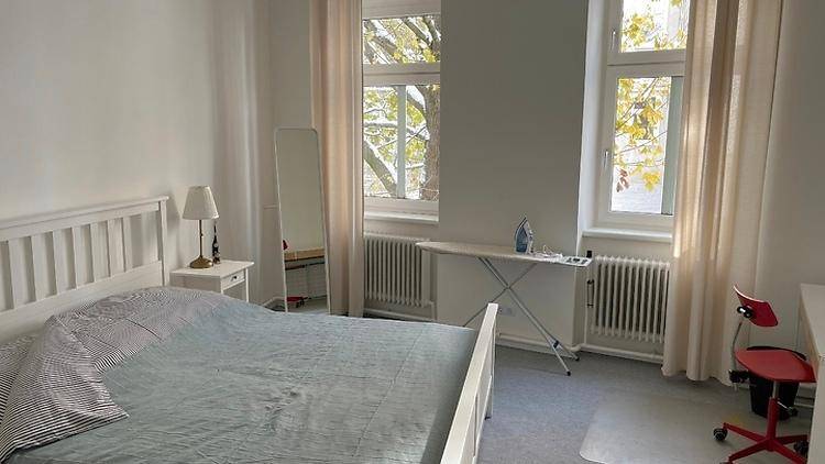 Helles Schlafzimmer mit weißem Bettrahmen, zwei großen Fenstern und einem Bügelbrett.