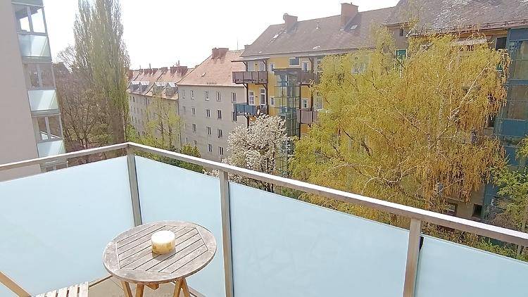 Kleiner Balkon mit Holztisch und Blick auf die umliegenden Gebäude und Bäume.