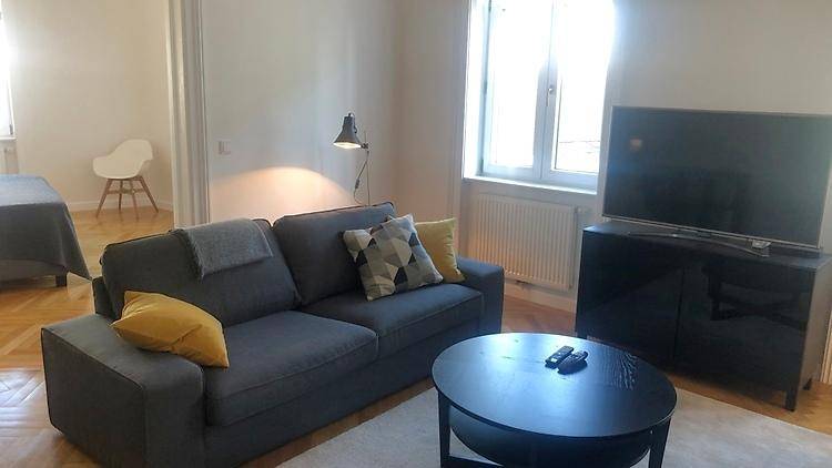 Gemütliches Wohnzimmer mit grauem Sofa, Couchtisch und Fernseher, mit Blick auf ein weiteres Zimmer.