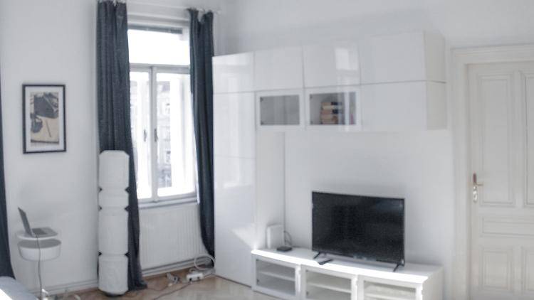 Wohnbereich mit Fernseher, weißen Hängeschränken und einem Fenster mit dunklen Vorhängen, hell und funktional.