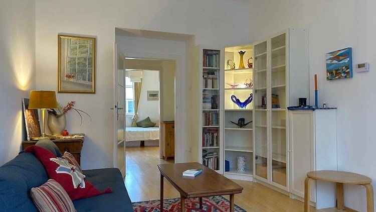 Wohnzimmer mit blauem Sofa, Holztisch und beleuchtetem Bücherregal, das zu einem Schlafzimmer führt.