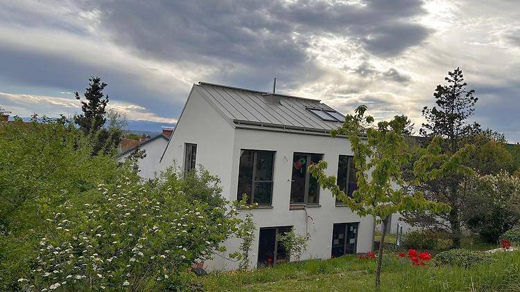 Modernes weißes Haus mit grauem Satteldach, umgeben von üppiger grüner Vegetation.
