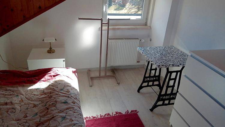 Helles Schlafzimmer mit Bett, Fenster, Heizkörper und einem Kleiderständer.