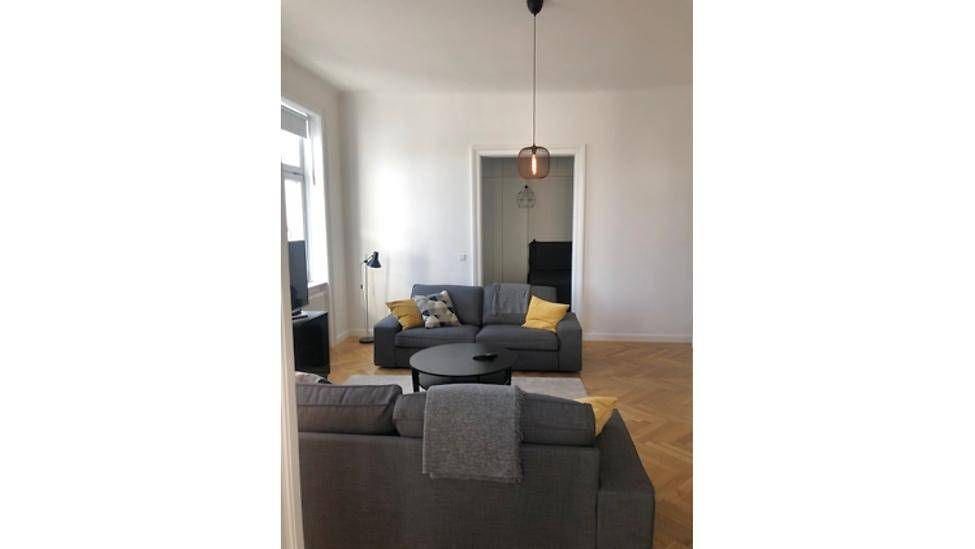 Geräumiges Wohnzimmer mit Parkettboden und moderner Hängelampe, offen zu einem weiteren Raum.