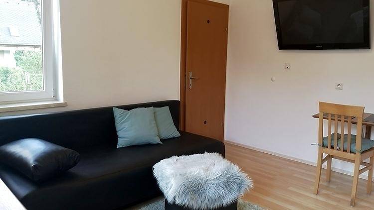 Heller Wohnbereich mit Sofa, Wandfernseher und einem kleinen Esstisch.