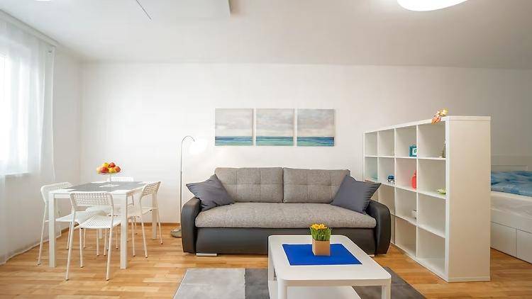 Helles Wohnzimmer mit Sofa, Esstisch und Regal, Teil eines Studio-Apartments mit Schlafbereich.