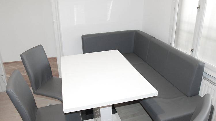 Gemütlicher Essbereich mit weißem Tisch, grauer Eckbank und passenden Stühlen, hell durch ein Fenster.