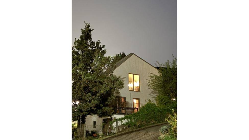 Hausfassade bei Nacht mit beleuchteten Fenstern und umgebenden Bäumen auf einem Hanggrundstück.