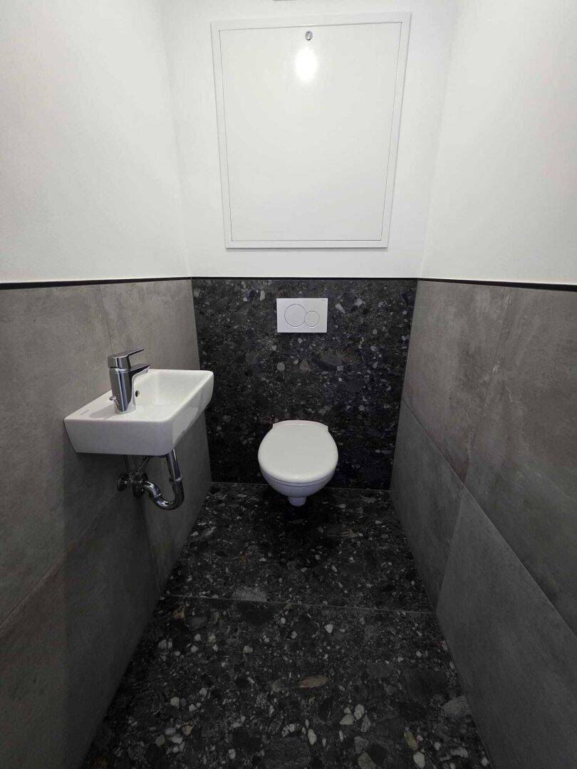 Kleines Gäste-WC mit Wand-WC, Waschbecken und dunklen Fliesen.