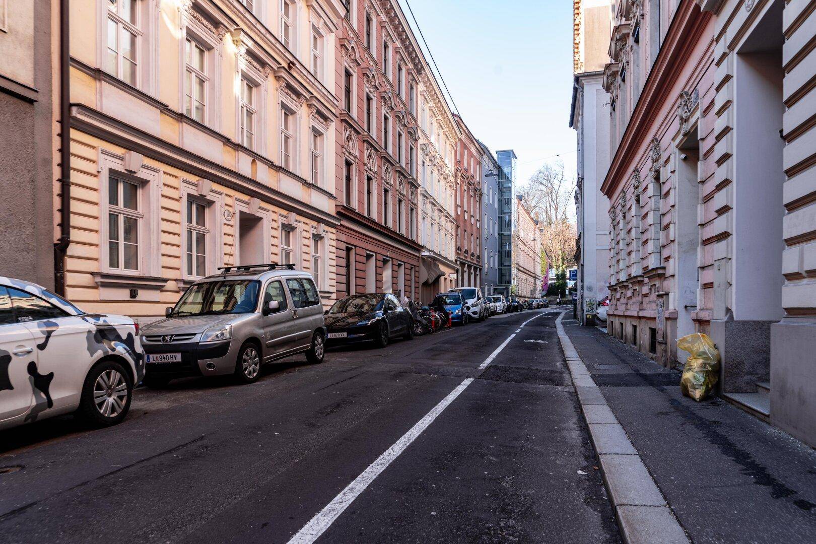 Ruhige Stadtstraße mit parkenden Autos und traditionellen Gebäudefassaden unter blauem Himmel, typisch für die Gegend.
