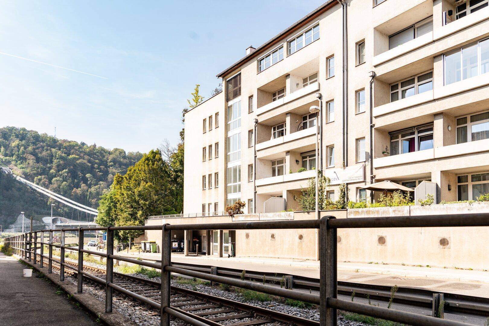 Modernes Mehrfamilienhaus mit Balkonen und großen Fenstern, gelegen an Bahngleisen und einem bewaldeten Hügel.