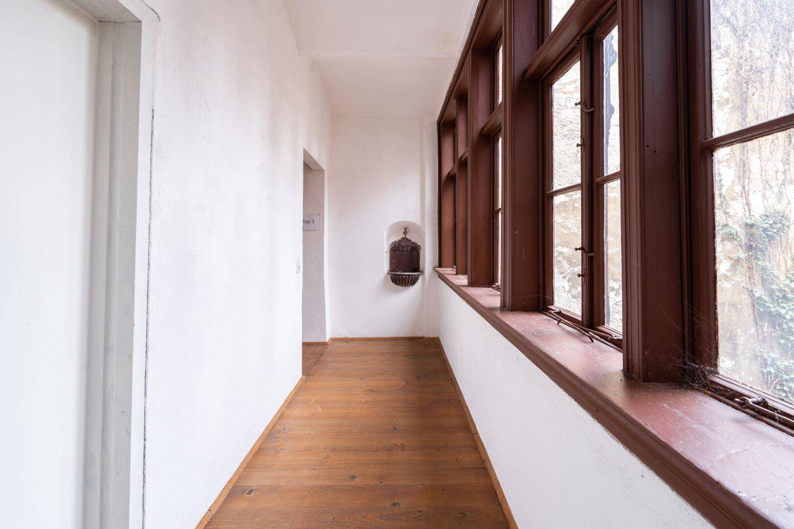 Heller Flur mit Holzdielenboden und großen, braunen Holzfenstern mit Blick ins Grüne.
