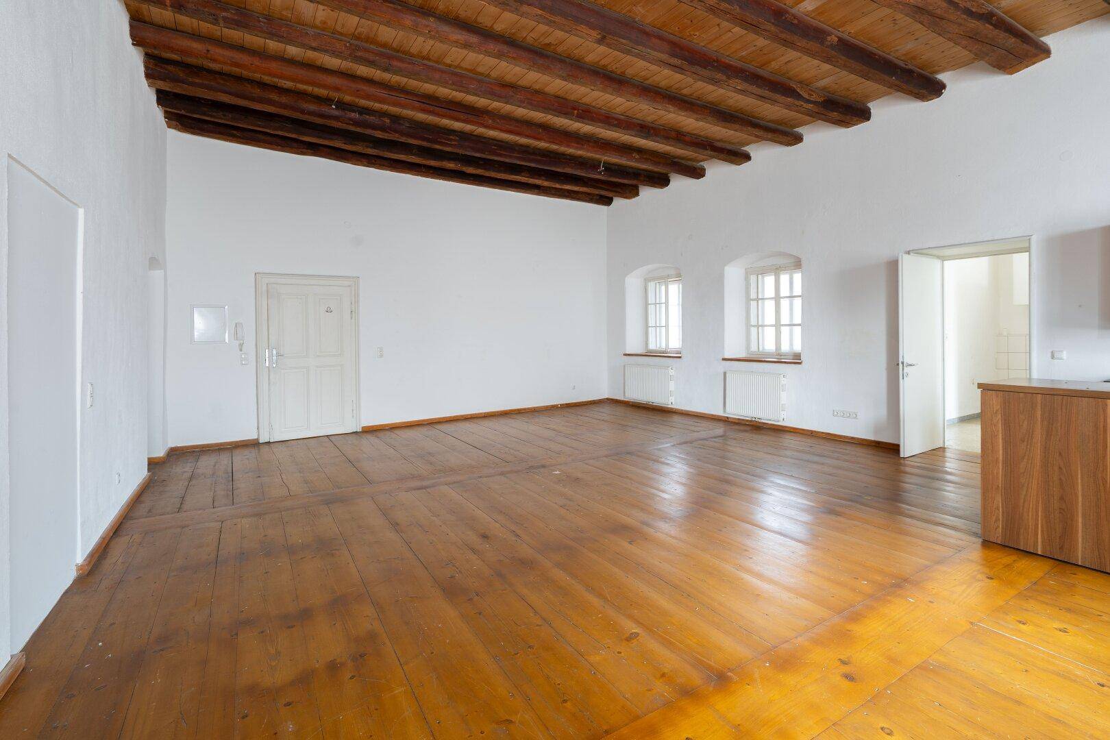 Geräumiger, leerer Raum mit Holzdielenboden, freiliegenden Holzbalken an der Decke und zwei Fenstern.