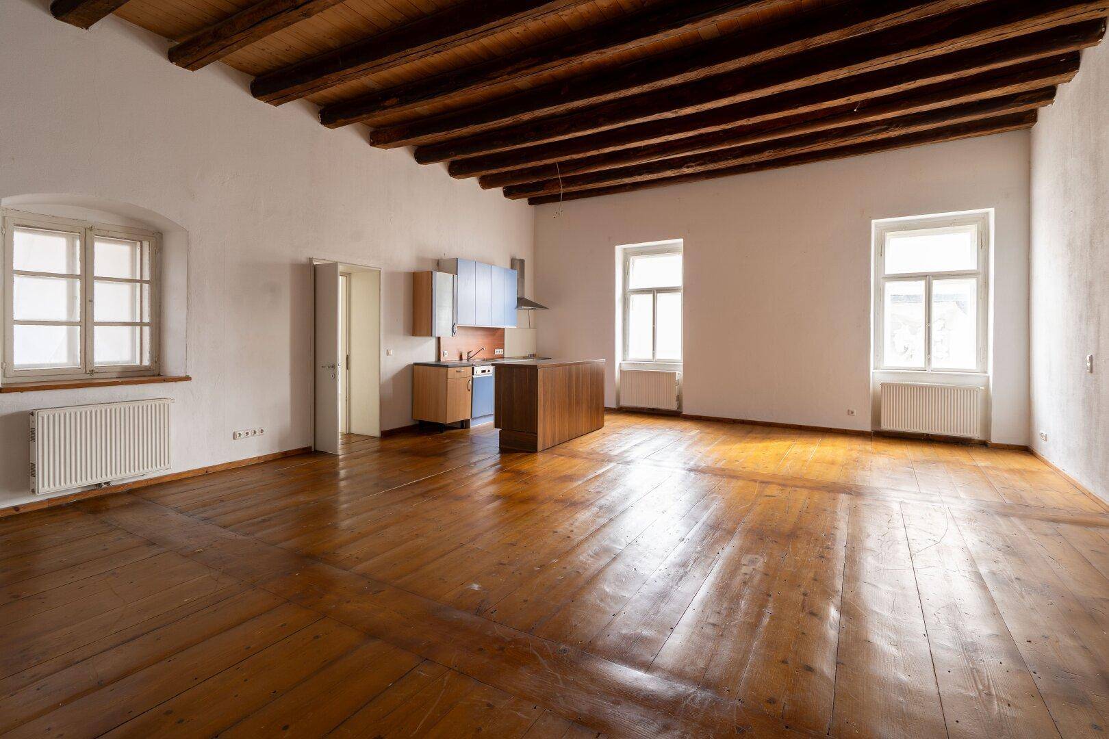 Großer Wohnbereich mit Holzdielenboden, sichtbaren Deckenbalken und einer kleinen Küchenzeile.