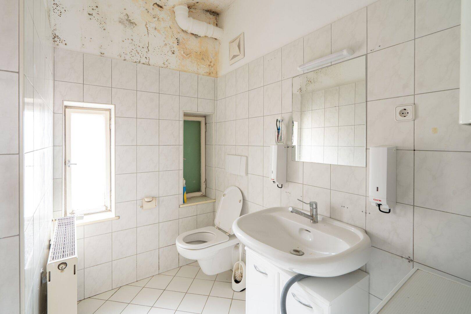 Badezimmer mit Fliesenboden, Waschbecken, Toilette und Fenster, zeigt deutliche Gebrauchsspuren.