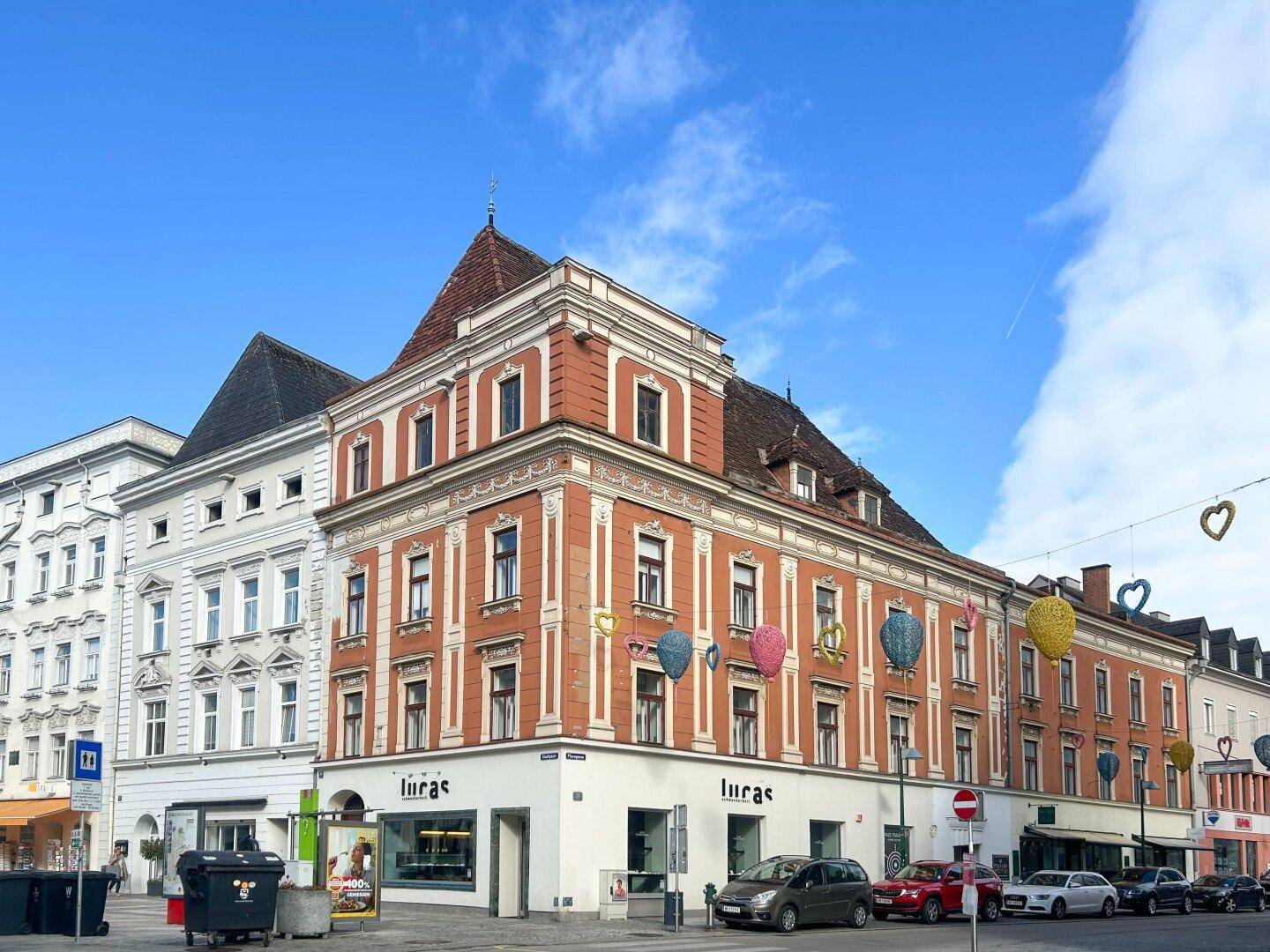 Eckgebäude mit markanter orangeroter Fassade und Geschäften im Erdgeschoss unter blauem Himmel.