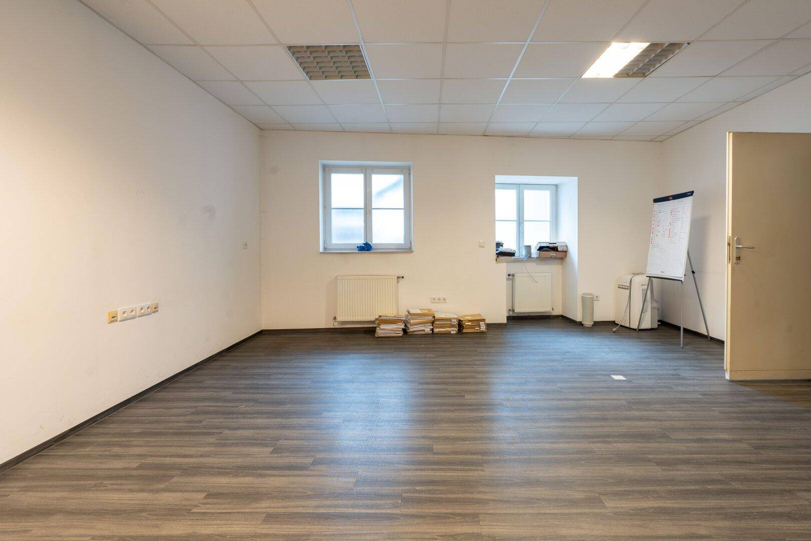 Geräumiger, leerer Büroraum mit Laminatboden, weißen Wänden und Rasterdeckenbeleuchtung, bereit zur Nutzung.