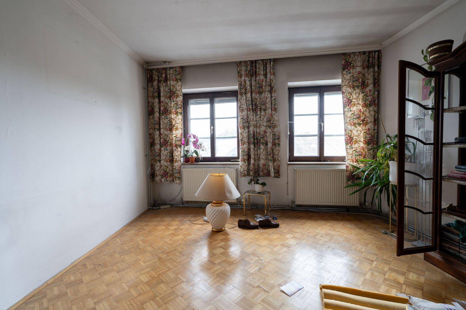 Gemütliches Wohnzimmer mit Parkettboden, zwei Fenstern und floralen Vorhängen, hell und einladend.
