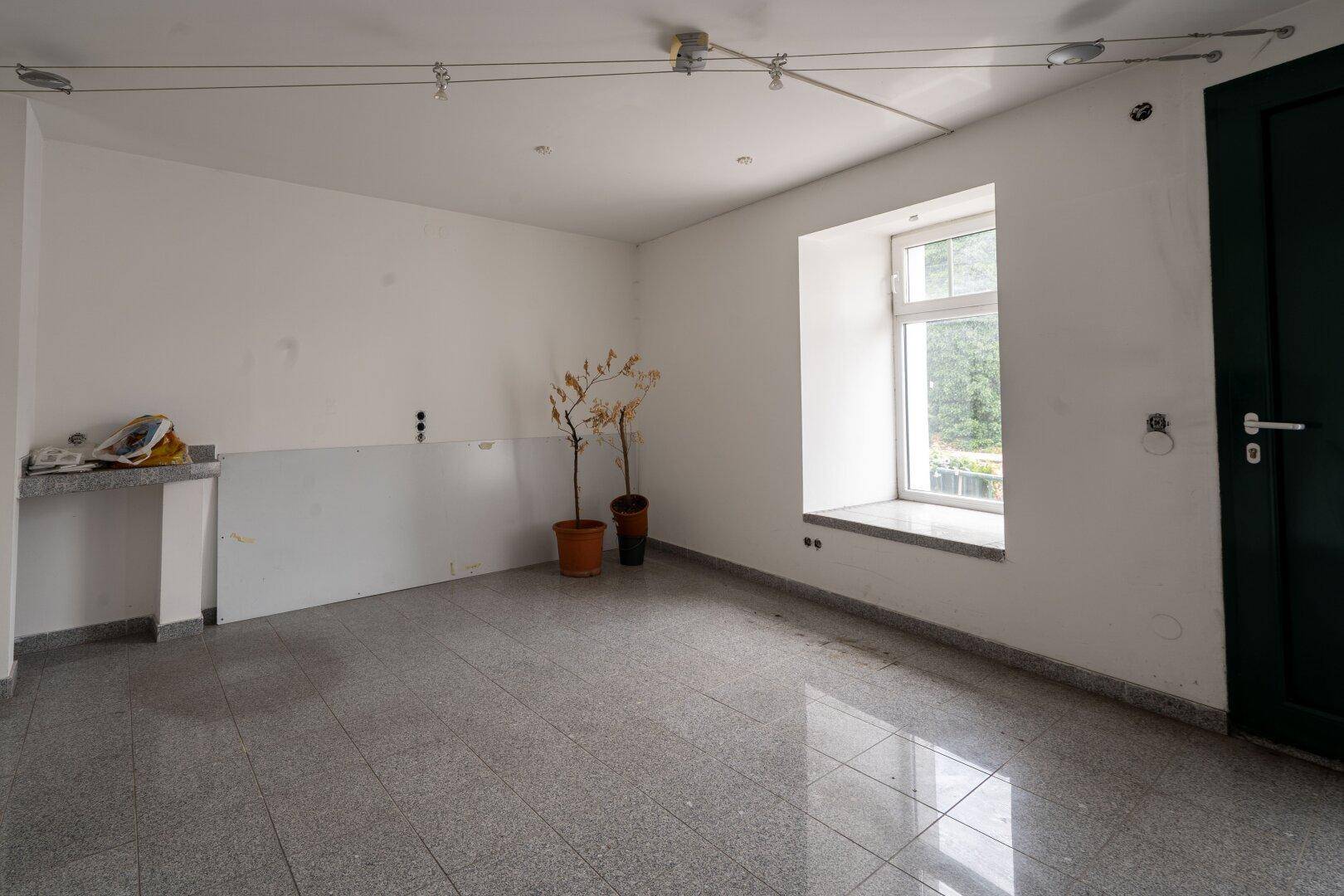 Leerer Raum mit Fliesenboden und einem Fenster, das Tageslicht hereinlässt, in neutralen Farben gehalten.