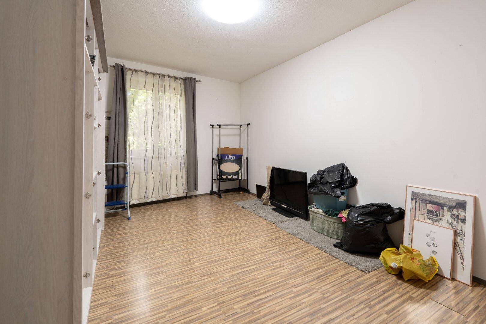 Einfaches Zimmer mit Laminatboden und einem Fenster, das viel Tageslicht hereinlässt, in neutralen Farben.