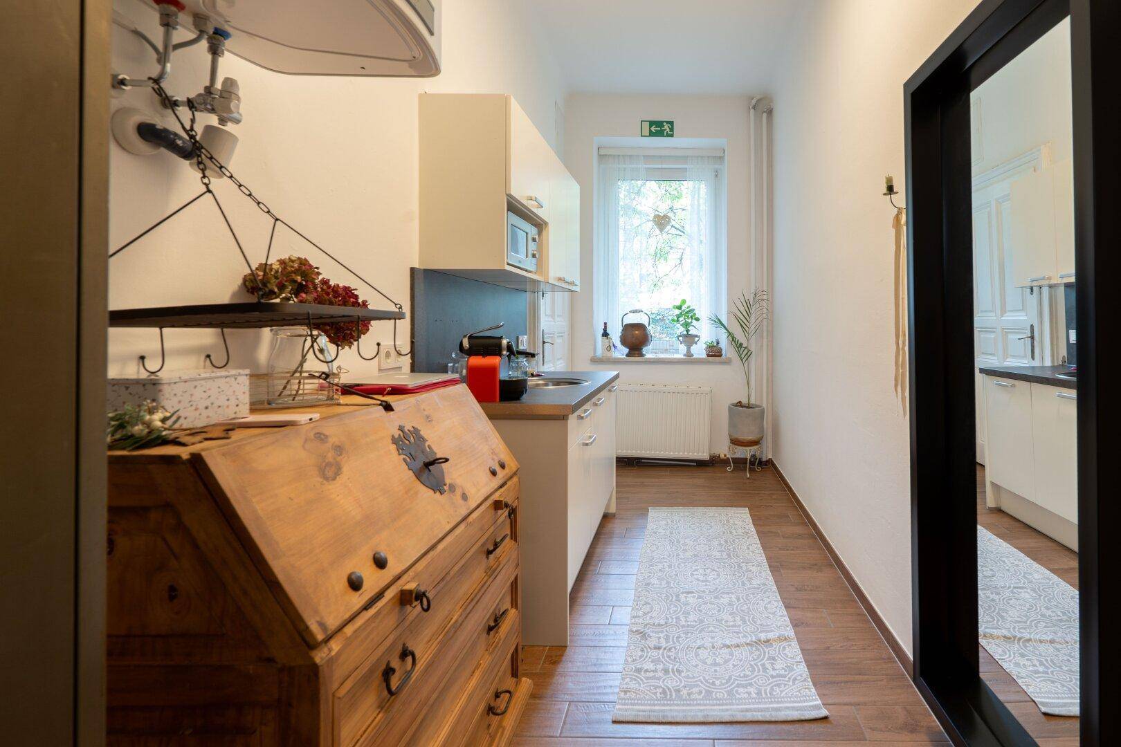 Schmale Küchenzeile mit weißen Schränken, Mikrowelle und Fenster, ergänzt durch einen antiken Holzschrank und Fliesenboden.