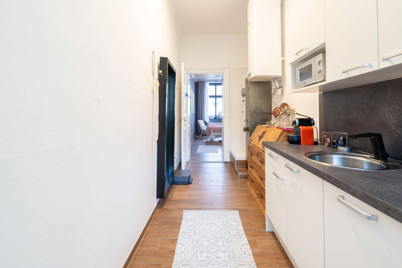 Funktionale Küchenzeile mit weißen Oberschränken, Spüle und Mikrowelle, führt zu einem weiteren Raum mit Holzboden.
