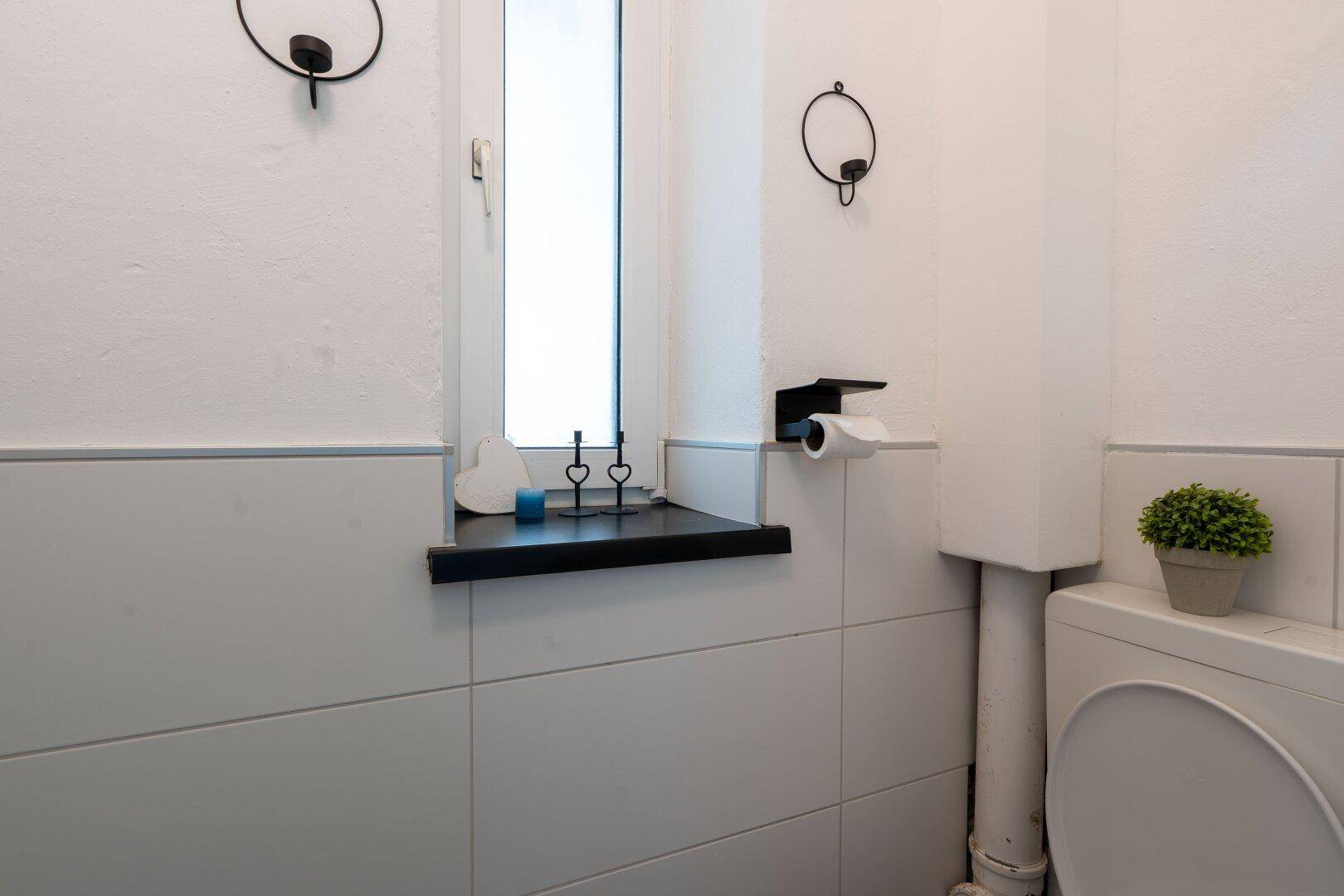 Helles Badezimmer mit weißen Fliesen, Fenster und einfacher Toilette, funktional eingerichtet und gut beleuchtet.