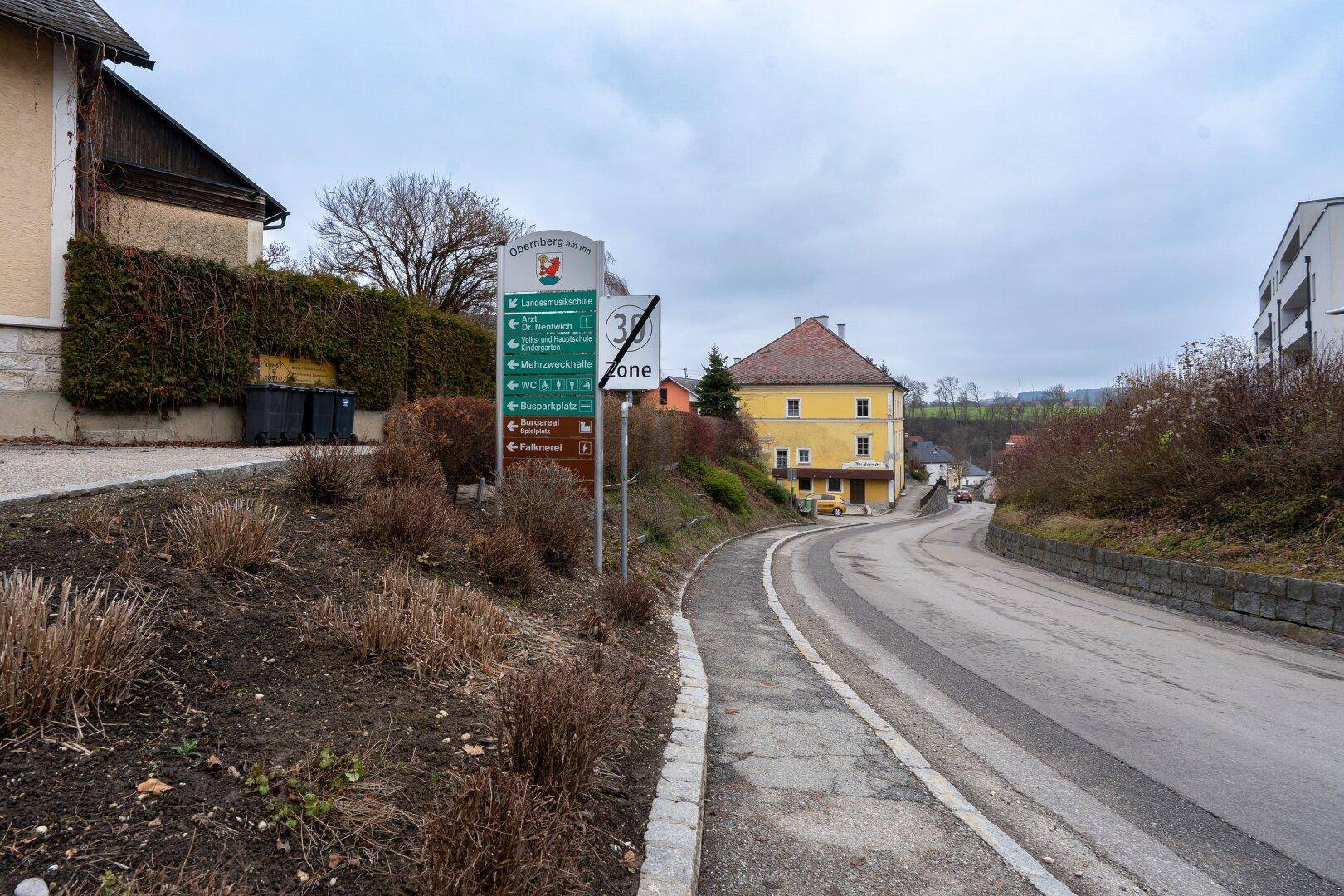 Ländliche Straße mit Wegweisern, umgeben von Vegetation und einem historischen Gebäude in der Ferne.