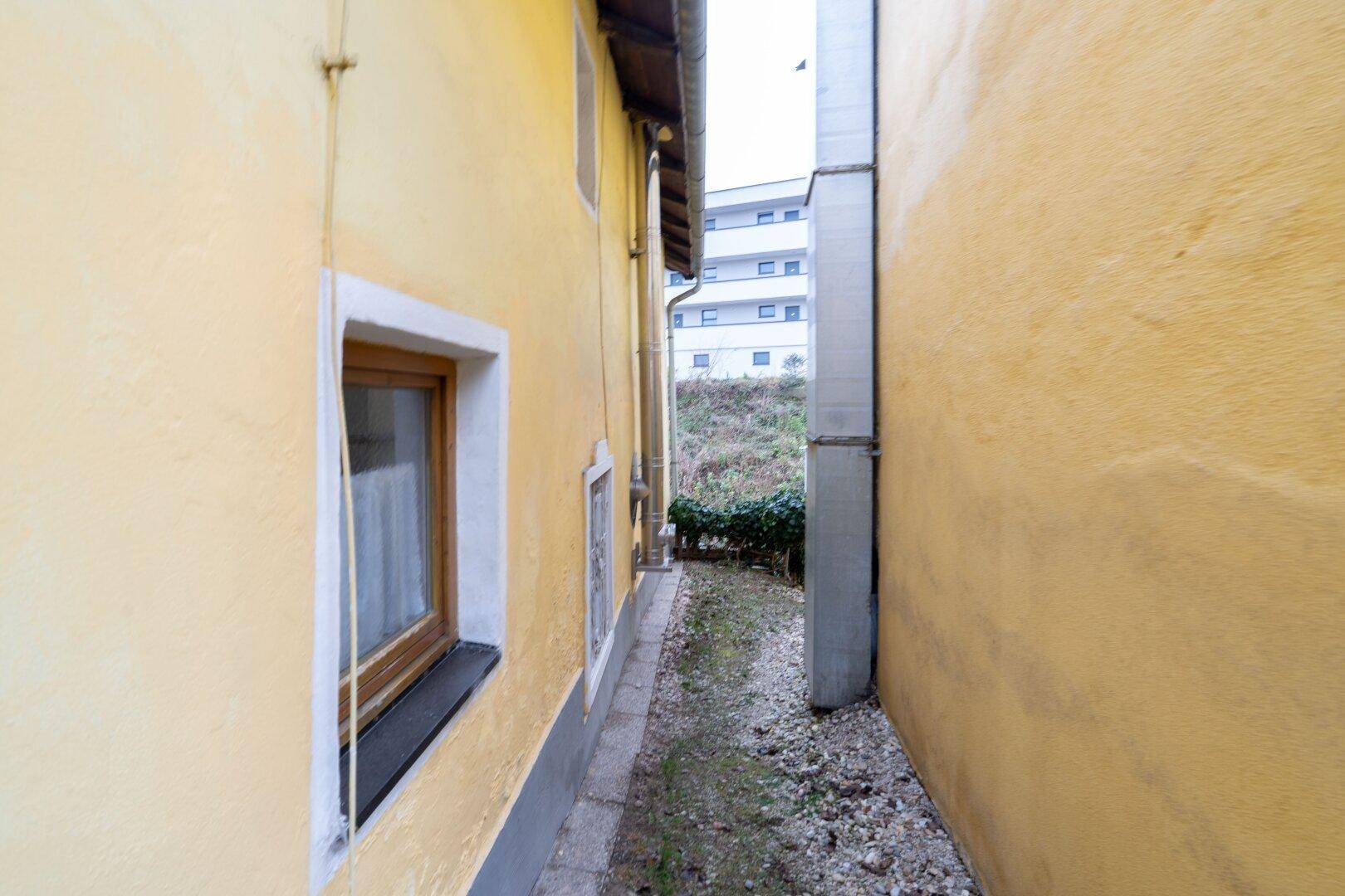 Schmaler Durchgang zwischen zwei Gebäuden mit gelber Fassade und Blick auf einen Neubau.