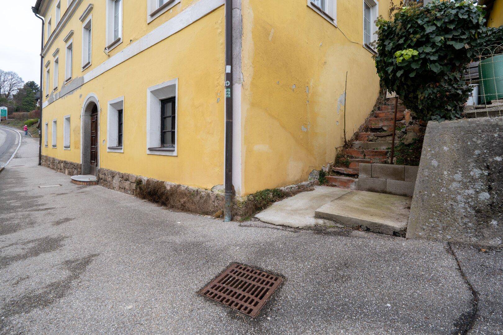 Ecke eines gelben Gebäudes mit Steinsockel, Fenstern und einer Außentreppe.